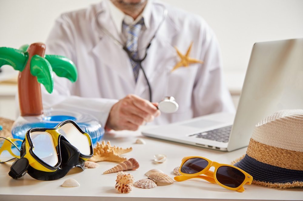 Medical and healthcare insurance for travel and summer holidays with a doctor seated at a laptop and goggles, sunglasses, sunhat and seashells in the foreground Sick on vacation, too bad? (Payroll Belgium)